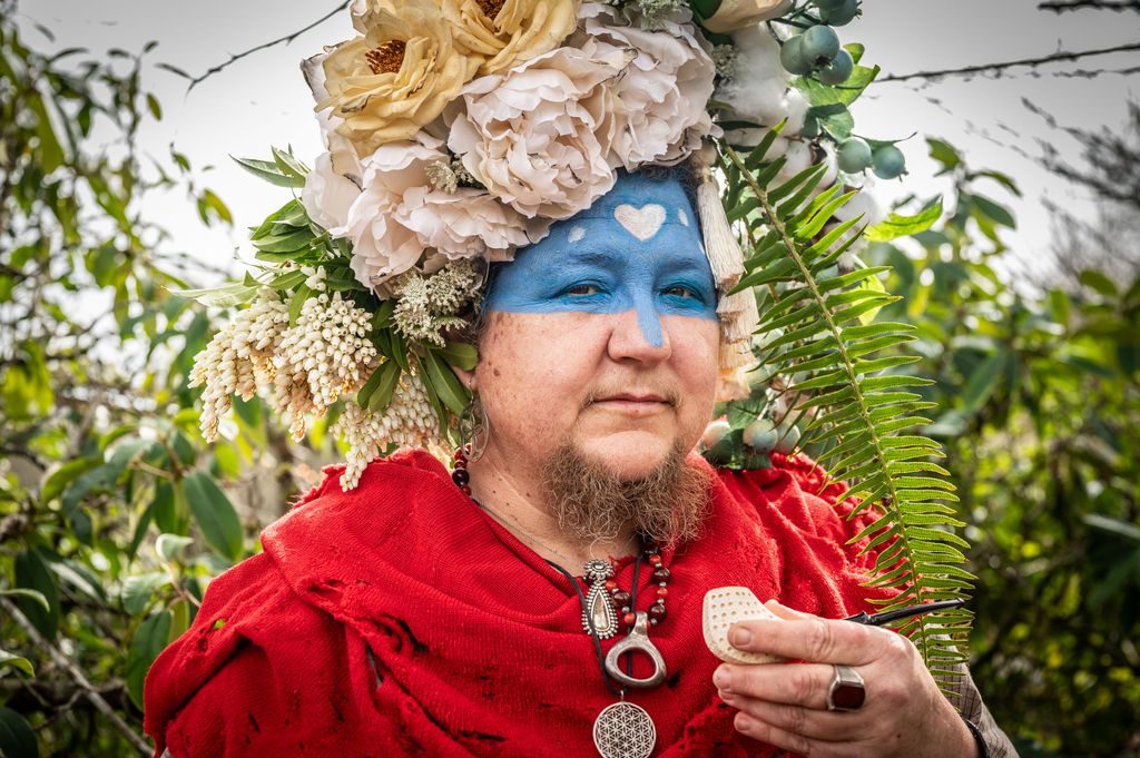 jennifer hawke, the artist, a white person with a beard and knowing eyes. the top half of their face is painted blue with a white heart, and they are wearing a huge elaborate flower crown, a red shawl, and several chunky pendants.